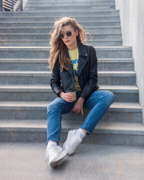 teen walk leisure. young blonde girl in jeans and black leather jacket sits casual on the gray street stairs background with coffee in hand and looks down in sunglasses. lifestyle concept, free space