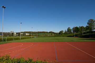 School empty soccer field near a forest in the hills. Daytime, blue sky, trees in the background, flood lights, no people
