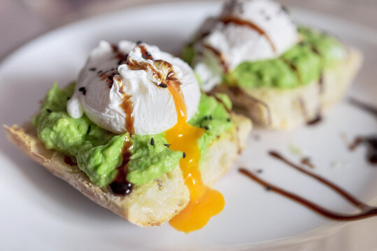 Poached egg on a slice of toast bread, with guacamole and dripping yolk, on a white plate