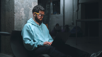 Male IT developer working with software and website at night on a laptop during the war in a dark bomb shelter