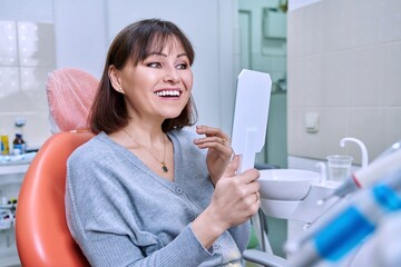 Smiling middle aged woman in dental chair with mirror looking at her teeth