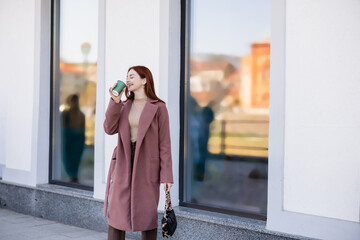 Fototapeta premium smiling redhead woman with handbag drinking coffee to go on urban street.