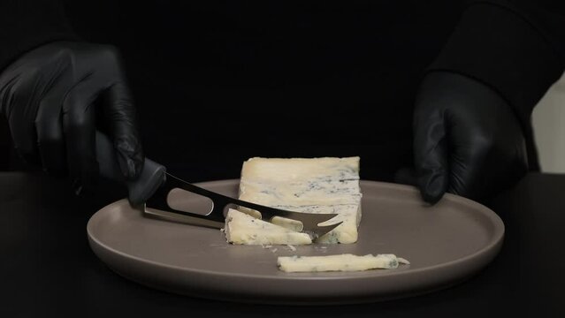 Pieces of blue cheese with knife on plate. Man's hands in black gloves cutting dorblu cheese. Cutting delicious cheese on black background. Man is chopping cheese