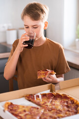 Blond teenager drinks cola and eats pizza in the kitchen at home. Portrait of a cute teenager