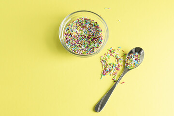 Confectionery multi-colored sprinkling in a transparent vase on a yellow table. A teaspoon on the...