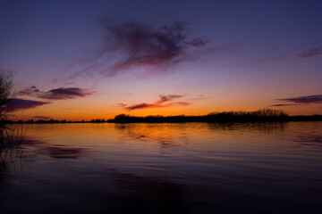Orange purple and violet sunset on river with dark colorful clouds in sky with trees reflection in water