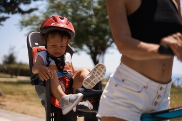 Portrait of little toddler boy with security helmet on the head sitting in bike seat and his mother with bicycle. Safe and child protection concept. Family and weekend activity trip