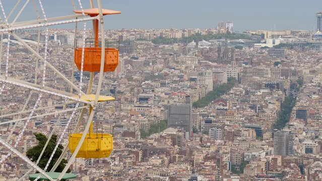 Empty gondolas of carousel wheel move against dense build city seen from height, fine telephoto perspective of Barcelona. Amusement ride at top of Tibidabo hill