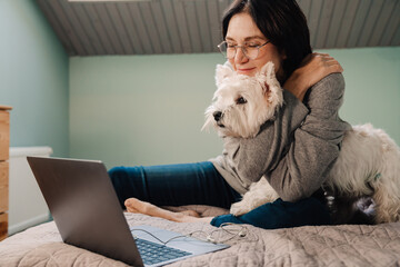 White mature woman using laptop while sitting with dog on bed