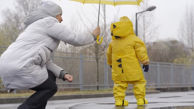 Mother with small cute child in yellow waterproof rain pants raincoat and rubber boots for kids walking outdoors in cold season, one year old happy baby boy on a rainy day. High quality 4k footage