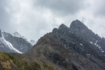 Obraz premium Atmospheric mountain landscape with large pointy pinnacle under gray cloudy sky. Gloomy scenery with high pointed mountain peak with sharp rocks in low clouds. Huge rocky peaked top in rainy weather.