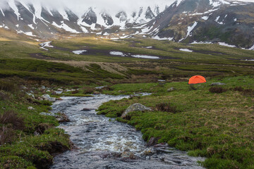 Orange tent near clear water stream with view to mountain range among low rainy clouds. Dramatic...