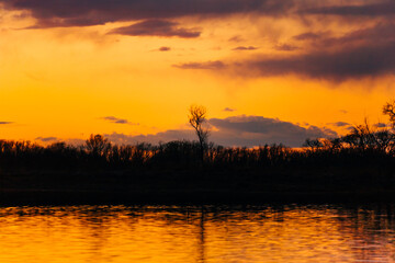 Fototapeta na wymiar Orange purple and violet sunset on river with dark colorful clouds in sky with trees reflection in water 