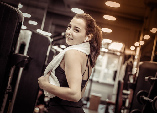 Portrait of young cheerful fit woman with towel on shoulders in modern gym