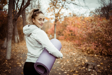 Attractive fit woman with yoga mat goes to workout in the autumn forest or park