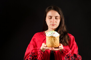 A young woman with her eyes closed holds an Easter cake with a lit candle on a black background, a place for text