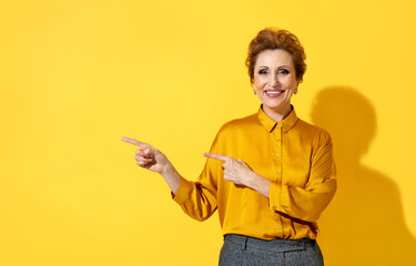 Smiling woman points away with both forefingers, shows on copy space for your text. Photo of attractive elderly woman in yellow shirt on yellow background