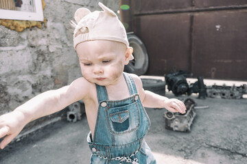 one year old baby in overalls with dirty face stands and waving his arms