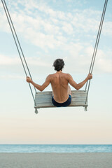 Young man traveler enjoying his summer vacation on swing by the sea. Swinging on the beach. Back...