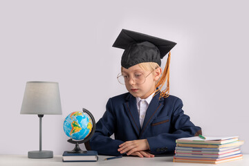 Portrait of boy in school uniform and students hat at table next to notebooks and globe. Education in junior school abroad.