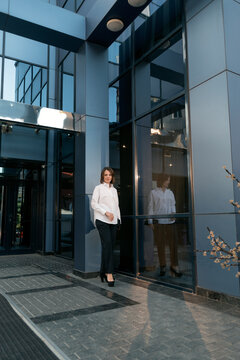 Full-length Portrait Of Young Business Woman In Formal Attire Against The Backdrop Of Modern Building With Glass Facade.