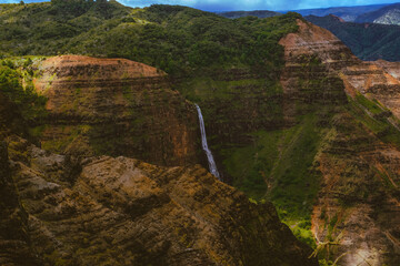 Waimea Canyon on the Hawaiian Island of Kauai, with red rocks and bright green foliage, dark gloomy clouds, bright blue sun, and long beautiful waterfalls 