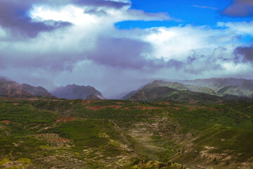 Waimea Canyon on the Hawaiian Island of Kauai, with red rocks and bright green foliage, dark gloomy clouds, bright blue sun, and long beautiful waterfalls 