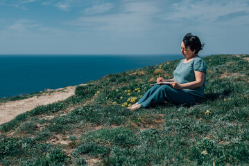 adult woman thoughtfully writes plans diary on a sunny day on a mountain against backdrop of the sea. pensioner sits on the grass barefoot in summer, spring, with a notebook and a pen