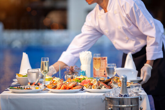 Waiter in white uniform serve of international food menu, shrimps, crab, seafood, champagne, wine on outdoor table at ceremony wedding party or hotel yard garden event. Dinner celebrates Fest concept
