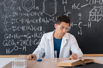 Young male chemistry teacher on blackboard background with chemical formulas, male 20s sitting at a...