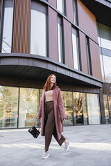 full length of pleased redhead woman in coat with handbag walking near modern building.