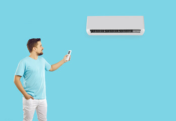 Happy young man using remote control turns on or adjusts air conditioner on light blue background. Caucasian man creates comfortable temperature during summer heat. Banner. Copy space. Advertising.