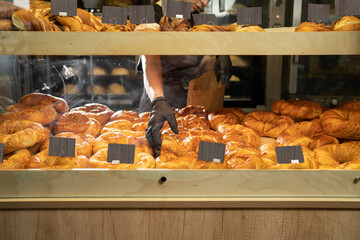 Freshly baked rolls behind a glass showcase of a store with price tags, seller's hand and delicious pastries for sale, many pastry products in the cafeteria, homemade baking