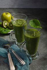 Two tall glasses of fresh home made green smoothies with ingrediends and spoons on green table.