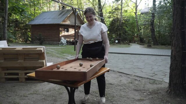 Happy caucasian woman playing vintage wooden game japanese billiards, culbuto in green yard of rural house. Lady tilts board, tries to hit target with wooden balls. Woman rejoices and claps her hands.