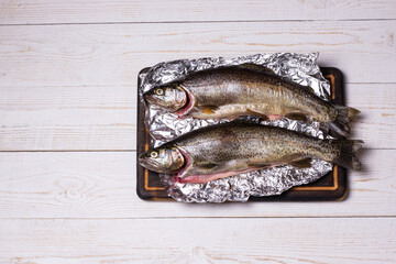 Raw trout fish on aluminum foil on wooden background, top view, flat lay image.