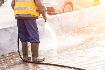 Man, worker in yellow orange vest and rubber boots washing sidewalk, road in sunlight. Cleaning street with hose with water