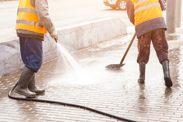 Two Man, worker in yellow orange vest and rubber boots washing sidewalk, road in sunlight. Cleaning...