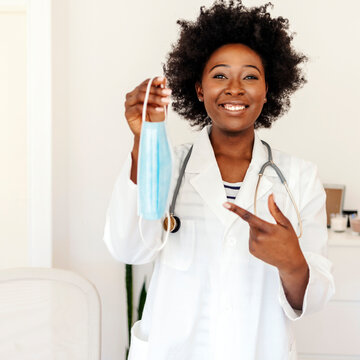 Portrait of young black African American woman in medical field, wearing a white coat and face mask, offering a face mask. Cropped portrait of an attractive female healthcare worker holding up a mask.