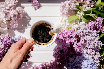 Obraz na płótnie Canvas White cup of fresh balck coffee on wooden white table with purple lilac branch. Morning routine. Coffee break concept. Bright spring flowers with green leaves and coffee flat lay. Top view.