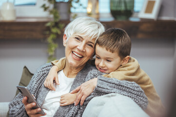 Grandma and grandson spend beautiful time together, they are happy and enjoy themselves. The child hugs her with a lot of love. Happy grandmother with her grandson in the house. - obrazy, fototapety, plakaty