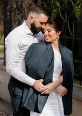 Front view of bearded man, wearing in white shirt, hugging woman and covering her shoulders by his suit jacket. Cute bride, dressed in man jacket while posing with groom outdoor