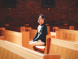 Man in black shirt praying in church with light sitting in the bench looking up