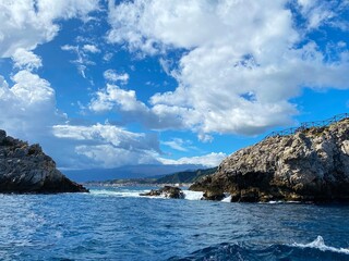 a picturesque view of the rocks in the sea against the backdrop of the coast of Sicily and white clouds on a blue sky on a sunny day