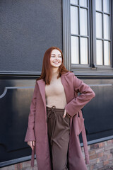 happy redhead woman in stylish coat standing with hand on hip near modern building.