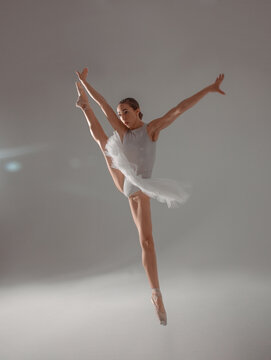 classic ballet trick. gymnast young dancer, fit girl ballerina jumps in white ballet tutu like white swan in cross twine in air near a white wall background in studio light. ballet concept, free space