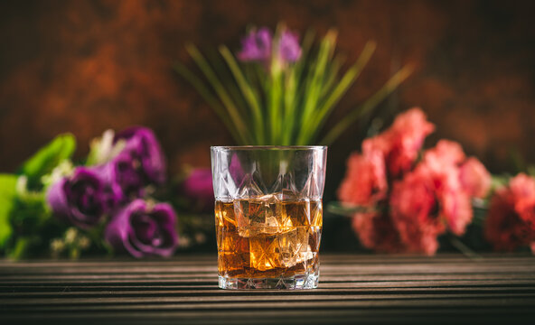 Glass of whiskey with ice on a wooden table with some color flowers. Alcoholic beverage ready to drink.