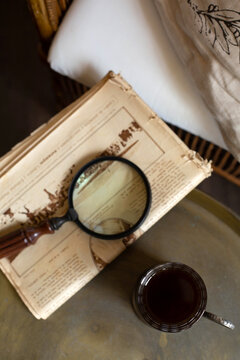 Glass of tea in an iron cup holder, a newspaper and a magnifying glass are on the table against the background of the interior in retro style. View from the top point. A vertical image.
