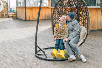 grandmother and granddaughter sat down to rest after a walk in a hanging rattan chair outdoors. an adult woman and a child are resting after a walk in autumn, spring on the street in outerwear
