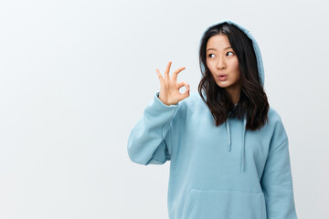 Friendly lovely cute Asian young female in blue hoodie sweatshirt show Okay sign gesture posing isolated on over white studio background. The best offer for ad. Fashion New Collection Ad concept